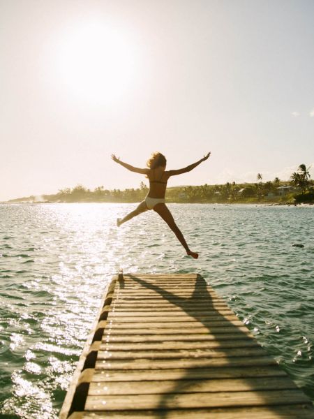 A person joyfully jumps off a dock into the water during sunset. The sun shines brightly over a scenic shoreline with palm trees.