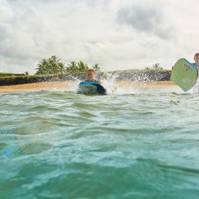 Two people are in the water at a beach, using bodyboards as waves splash around them under a cloudy sky.