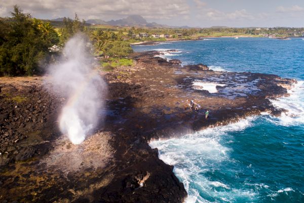 The image shows a coastal landscape with a blowhole spouting water and people nearby, against a backdrop of lush greenery and ocean waves.