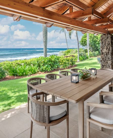A beachfront patio with a wooden dining table and chairs under a wooden roof, overlooking the ocean with lush greenery around.
