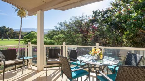 Outdoor patio with a glass dining table set for a meal, surrounded by chairs, overlooking a garden and mountains in the background.