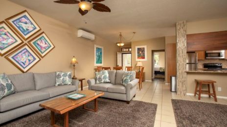 A cozy living room with gray sofas, a wooden coffee table, colorful artwork, a kitchen area, and a dining table in the background.