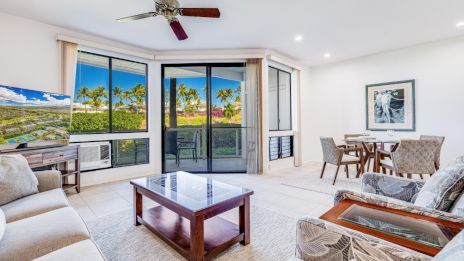 A bright living room with sofas, a coffee table, TV, dining area, large windows, and a sliding door showing a green outdoor view.