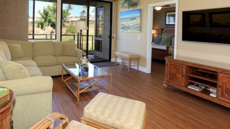 A cozy living room with a large TV, beige couch, glass coffee table, and wooden furniture. A sliding door leads to a balcony.