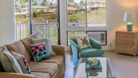 A cozy living room with a sofa, armchair, and glass table, featuring tropical decor. Large windows offer views of palm trees outside.