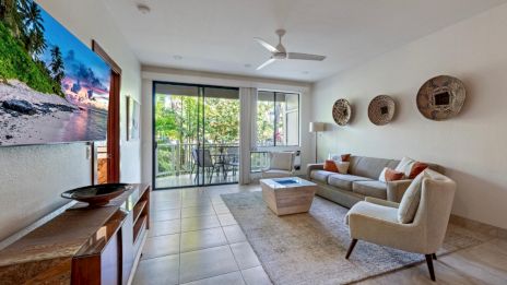 A modern living room with a couch, coffee table, and wall art. Large windows show a balcony and greenery outside.