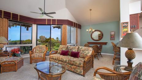 The image shows a cozy living room with wicker furniture, a glass table, large windows, floral patterns, and a bar area with a mirror.