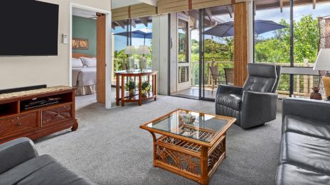 A living room with leather sofas, glass coffee table, TV, and a balcony view with outdoor seating, next to an open door to a bedroom.