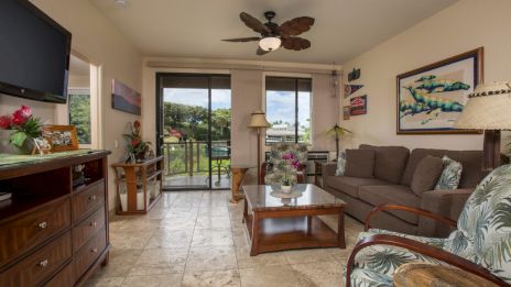 A cozy living room with a sofa, armchair, coffee table, and wall-mounted TV. Artwork and plants add decor; a glass door leads outside.