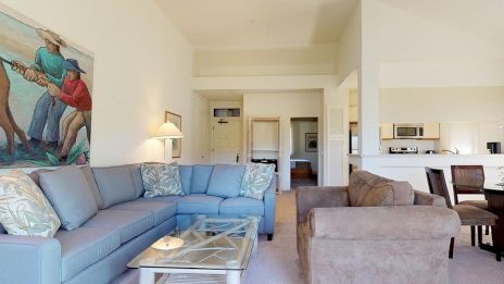 A cozy living room features a blue sectional sofa, glass coffee table, artwork, and a dining area near an open kitchen space.