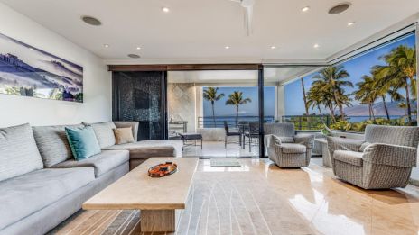 A modern living room with a grey couch, armchairs, and a coffee table; it features large windows with a view of palm trees and the ocean.