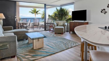 A modern living room with a sofa, armchair, TV, and glass coffee table opens to a balcony with ocean views and palm trees.