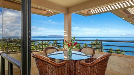 A balcony with a table, wicker chairs, and a vase of flowers overlooks a scenic ocean view with clear skies and palm trees.