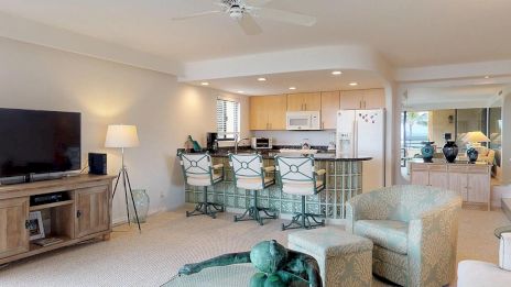 A cozy living room with a TV, comfy chairs, kitchen area with bar stools, and a unique green statue on the glass coffee table.