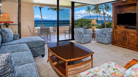 A cozy living room with a blue sofa, floral chair, TV, and a beautiful ocean view through glass doors leading to a balcony with seating.