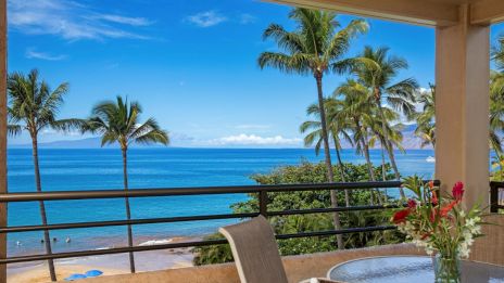 A patio overlooking a tropical beach with palm trees and ocean view; a table with flowers in the foreground completes the scene.