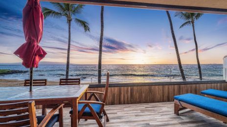 A beachfront patio with wooden furniture overlooks the ocean at sunset, framed by palm trees and a vibrant sky.