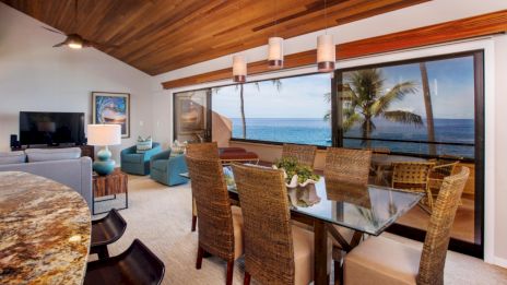 A modern dining and living room is shown with ocean views, large windows, and cozy seating under a wood ceiling.