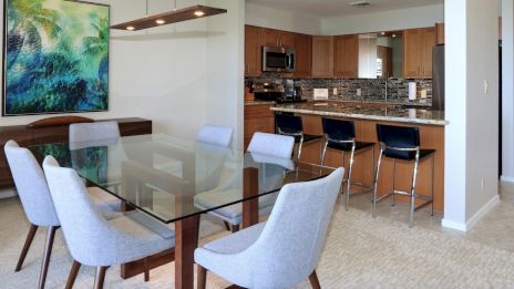 A modern dining area with a glass table, gray chairs, and a kitchen with a bar counter and stools, featuring a large abstract painting.