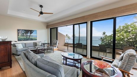 A spacious living room with large windows, ocean view, modern furniture, ceiling fan, and outdoor seating area visible outside.