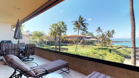 A patio with lounge chairs, a table, and a grill overlooks a scenic view of palm trees, houses, and the ocean on a sunny day.