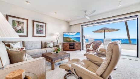 A modern living room features plush seating, framed art, and a TV, with a stunning ocean view through large open sliding doors.