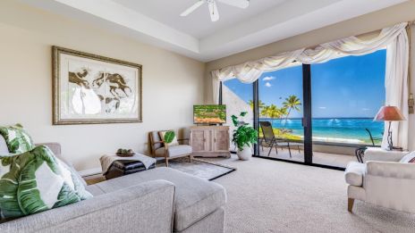A bright living room with modern furniture, a large window showing a beach view, and tropical decor touches throughout, creating a cozy atmosphere.
