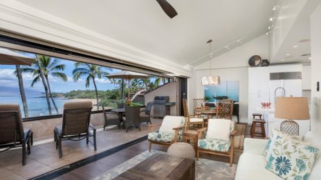 A spacious open-concept living area with beach views, featuring cozy furniture, a fan, and patio seating overlooking the ocean and palm trees.