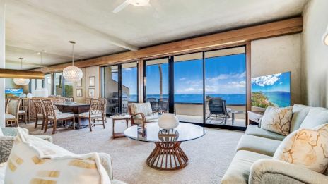 The image shows a spacious living room with large windows, comfortable seating, a round coffee table, and a stunning ocean view.