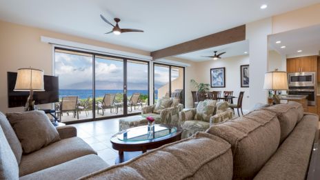 A spacious living room with modern furnishings, ocean view, large windows, ceiling fans, and a dining area in the background.