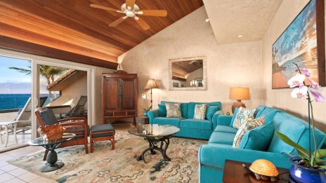 A coastal living room with turquoise sofas, wooden ceiling, a fan, and a view of the ocean through sliding glass doors.