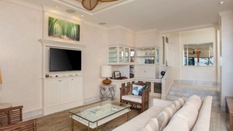 The image shows a living area with a white sofa, chairs, a TV, glass coffee table, and an open-plan office space in an elegant, neutral decor.