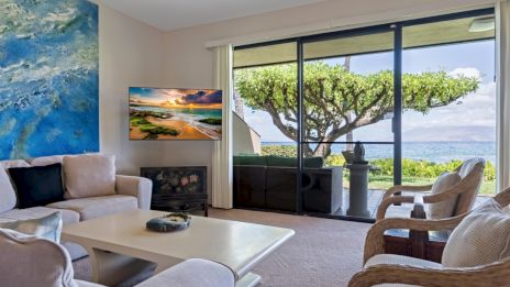 A cozy living room with beige furniture, abstract art, and a TV. Large windows reveal a lush view with a uniquely shaped tree and ocean.