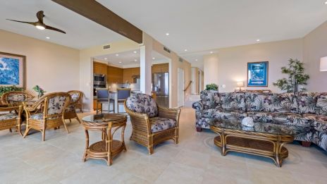 A spacious living room with wicker furniture, tile floor, ceiling fan, and art on walls, connected to dining and kitchen area.
