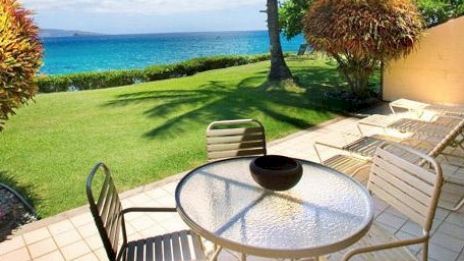 A patio with a glass table and chairs overlooks a lush lawn, palm trees, and a tranquil ocean view under a clear blue sky.