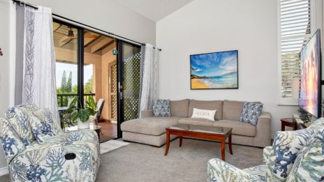 A cozy living room with floral armchairs, a beige sofa, TV, and balcony access. A ceiling fan and beach artwork complete the decor.