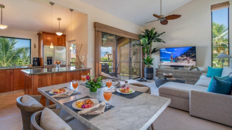 Open living space with a dining table, sofa, TV, and a kitchen; bright and airy with tropical plants and decor.