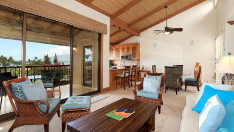 A cozy living room with wicker furniture, a wooden table, a view of a balcony, and a kitchen area in the background.