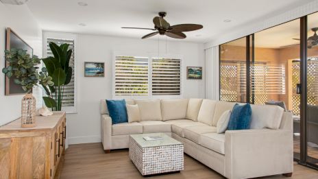 A modern living room with a beige sectional sofa, blue cushions, and a wicker coffee table, featuring large windows and a ceiling fan.