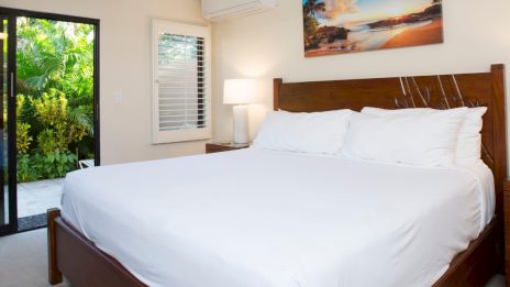 A bedroom with a large white bed, a landscape photo on the wall, a window, and a glass door leading to a leafy outdoor area.