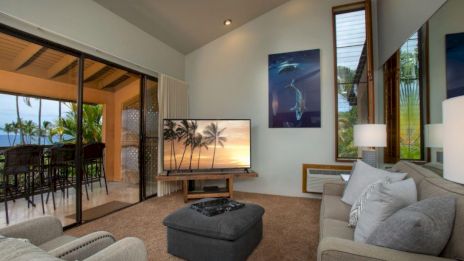 A cozy living room features a TV, sectional sofa, and sliding doors to a patio with bar stools. Ocean view and tropical decor complete the scene.