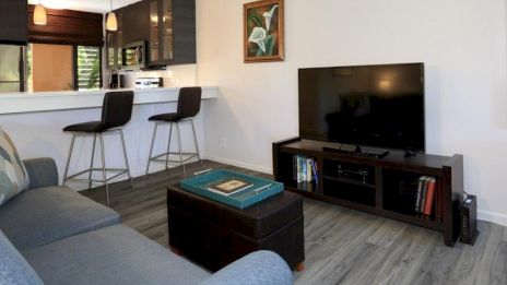 A cozy living area features a sofa, coffee table, TV stand with books, and view into the kitchen with bar stools and pendant lights.
