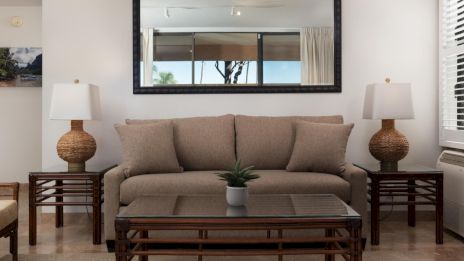 A cozy living room with a beige couch, glass coffee table, and two table lamps, accented by a large mirror on the wall.