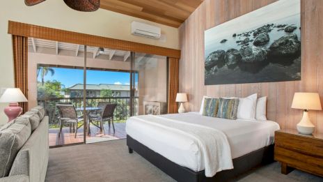 A cozy bedroom with a large bed, sofa, lamps, and a nature-themed wall art has a view of a balcony through sliding glass doors.