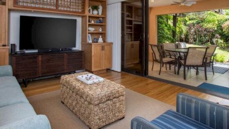 The image shows a cozy living room with a TV, wicker ottoman, blue sofa, and open sliding door to an outdoor patio with dining area.