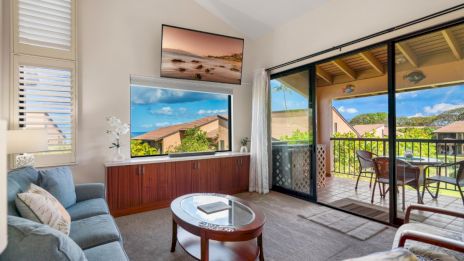 A cozy living room features a sofa, coffee table, wall-mounted TV, and a balcony with outdoor seating and scenic views.