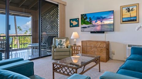 A cozy living room with teal sofas, a TV, and beach-themed decor. A balcony with palm trees is visible through sliding glass doors.