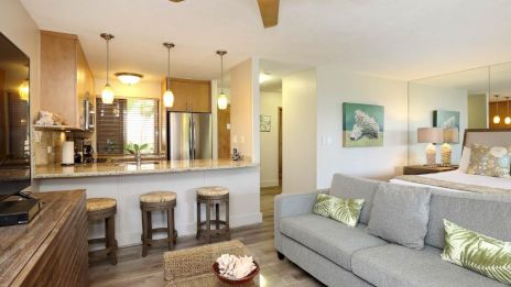 A cozy living room with a sofa, coffee table, and kitchen with bar stools and pendant lights in the background.