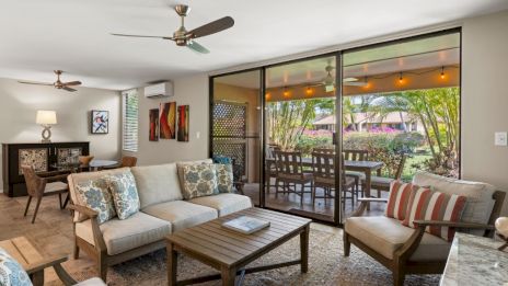 A cozy living area features a sofa, armchairs, and wooden table, with a view of a patio through large glass doors and tropical greenery.