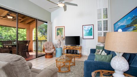 A cozy living room with a TV, comfortable seating, art on walls, and doors opening to a patio with outdoor dining furniture and nature views.